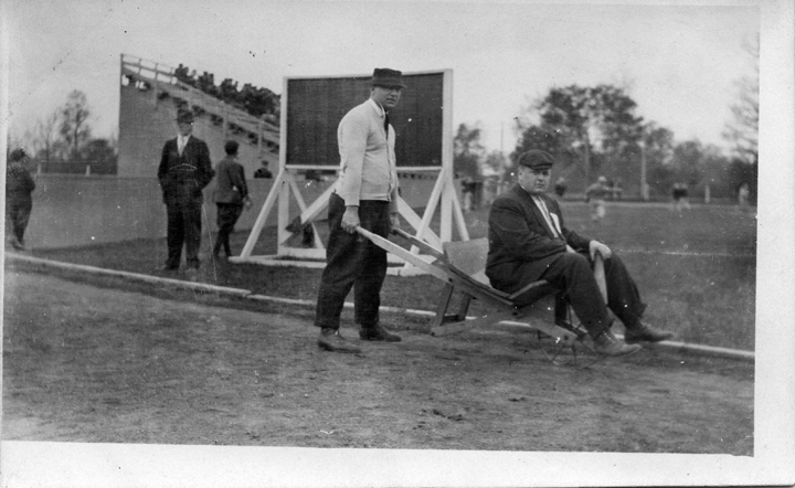 A man pushes a wheelbarrow while another man sits on it